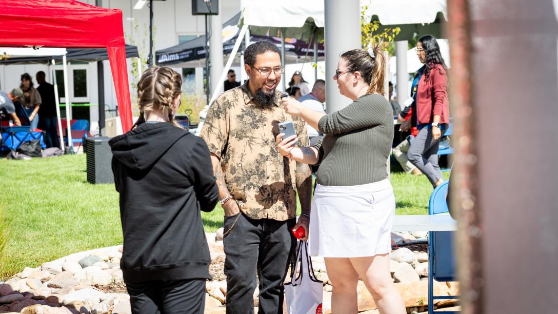 A student holds a microphone up to another student while showing them a picture on a phone. Another person is filming them with their phone while people in the background wander between tents at the annual Fort Lupton Fall In.
