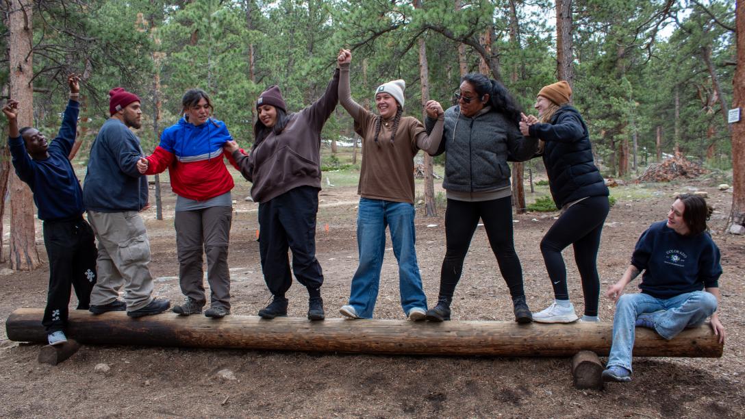 Student leaders standing on a log in a forest holding hands as they participate in a team building activity involving balance