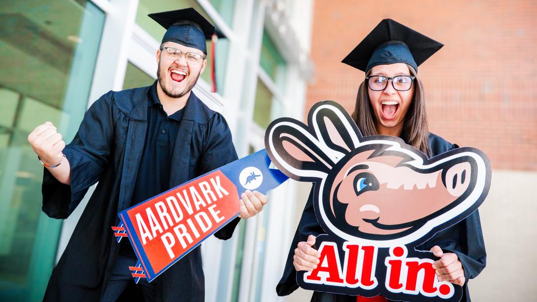Aims students in cap and gown with All in and Aardvark Pride signs