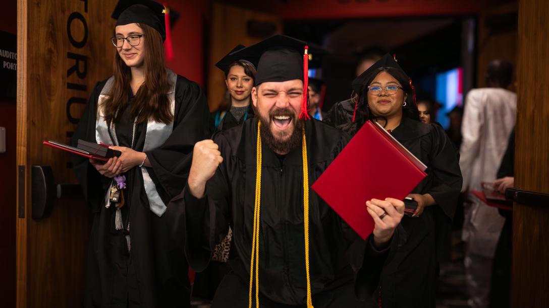 An Aims graduate looks at the camera with one hand raised in excitement and their diploma in the other hand