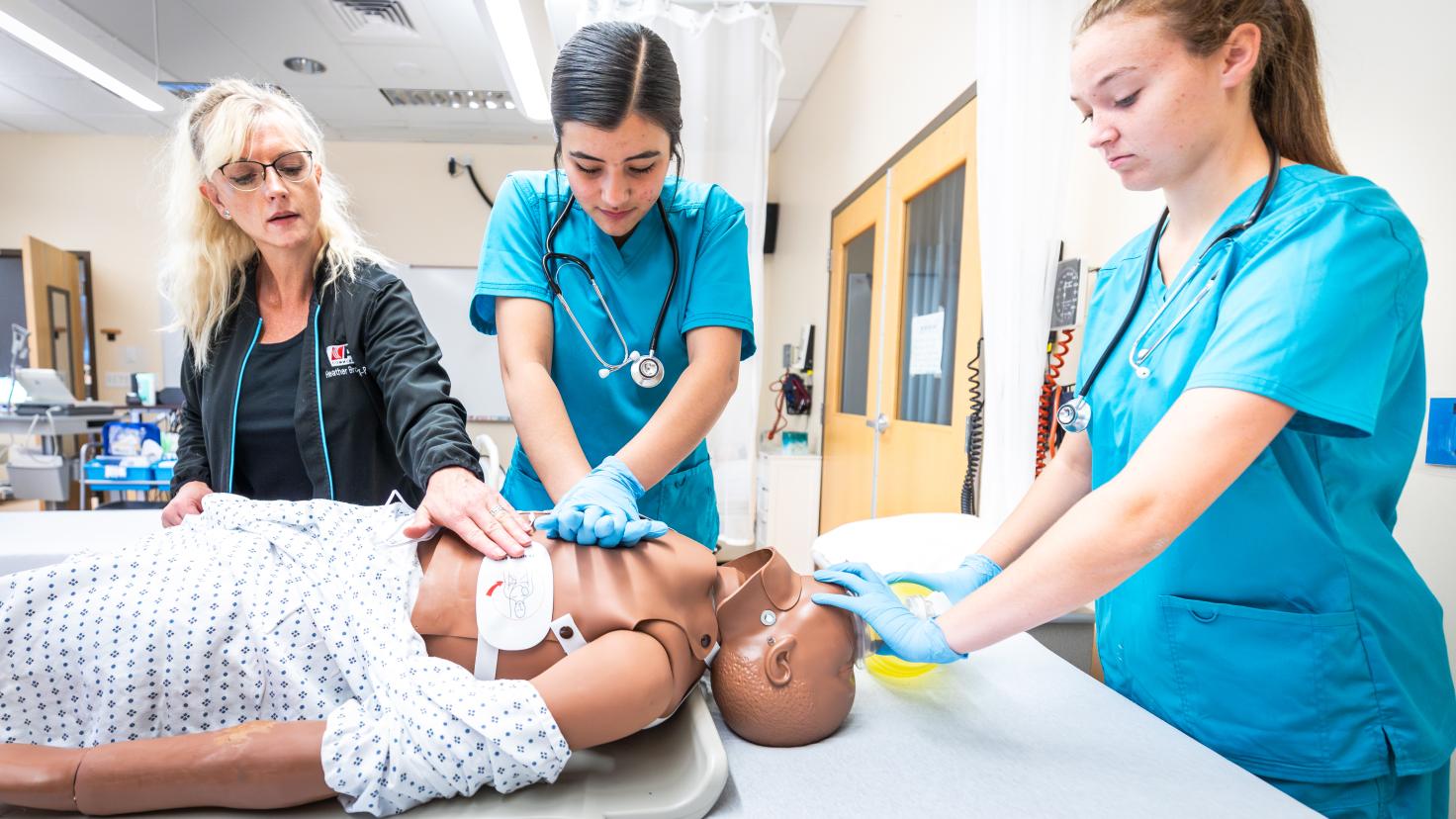 3 acute care nursing students in blue scrubs practice chest compressions on CPR doll