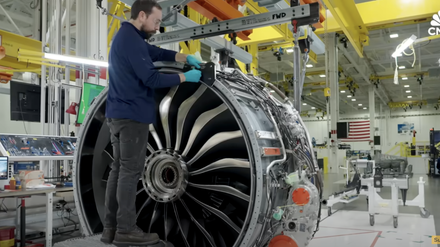 A technician in a blue shirt and gloves works on a large jet engine in a brightly lit aerospace facility