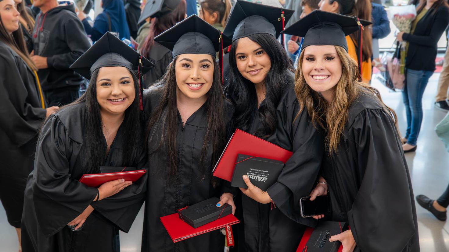 Four spring 2023 Aims graduates smiling wearing caps and gowns and holding diplomas. Other graduates and ceremony attendees are in the background.