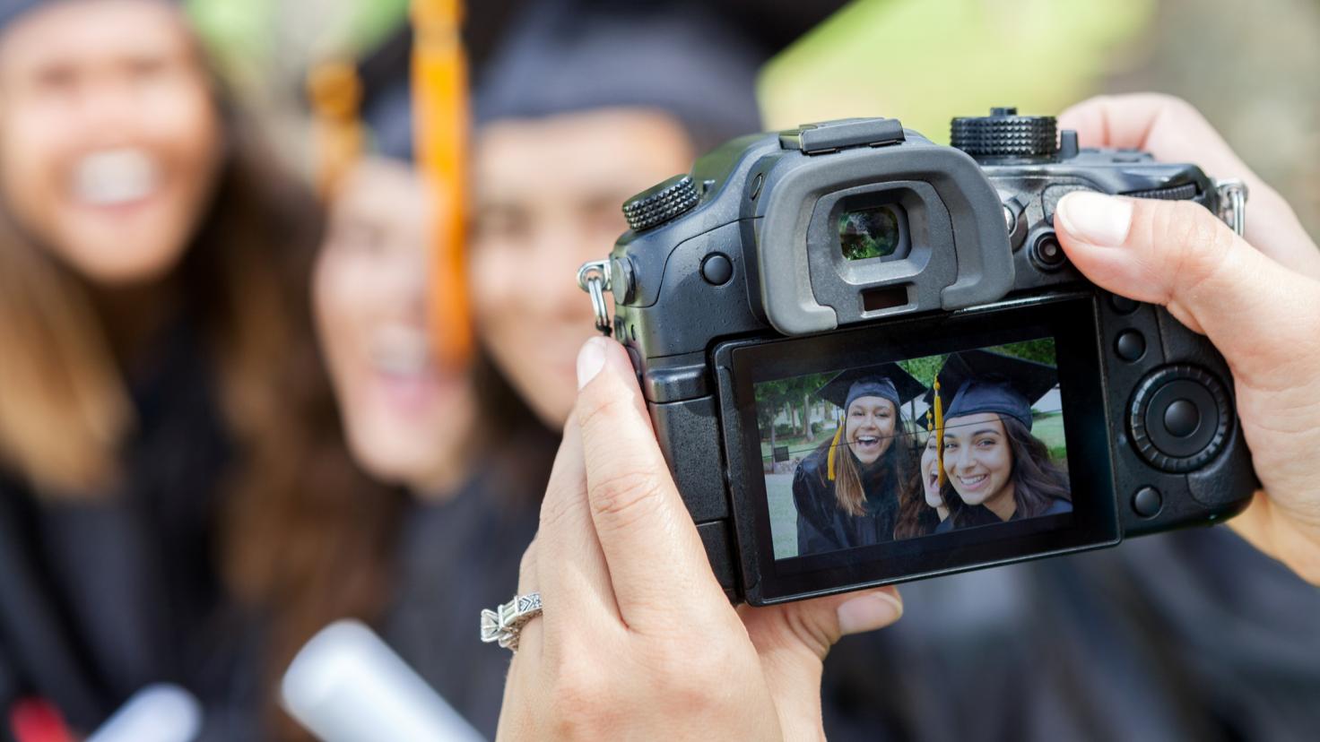 Female photographer takes a photo of three college graduates and friends. They are holding diplomas and smiling at the camera. Focus is on the camera in the foreground.