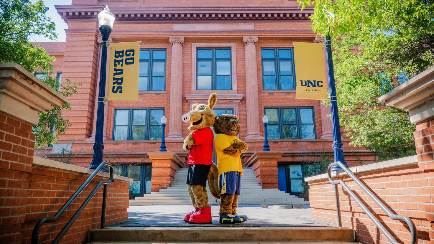 Aims and UNC mascots, Arty and Klawz standing in front of a UNC building back-to-back with arms crossed facing camera