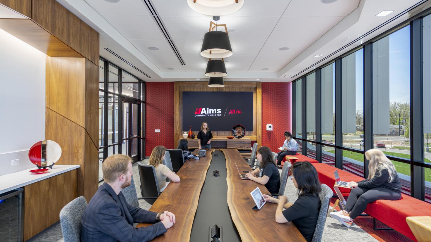Aims Executive Conference Room in the Welcome Center showing long wood table with people sitting at it and a wall of windows with a bench and a woman sitting on the bench using a laptop