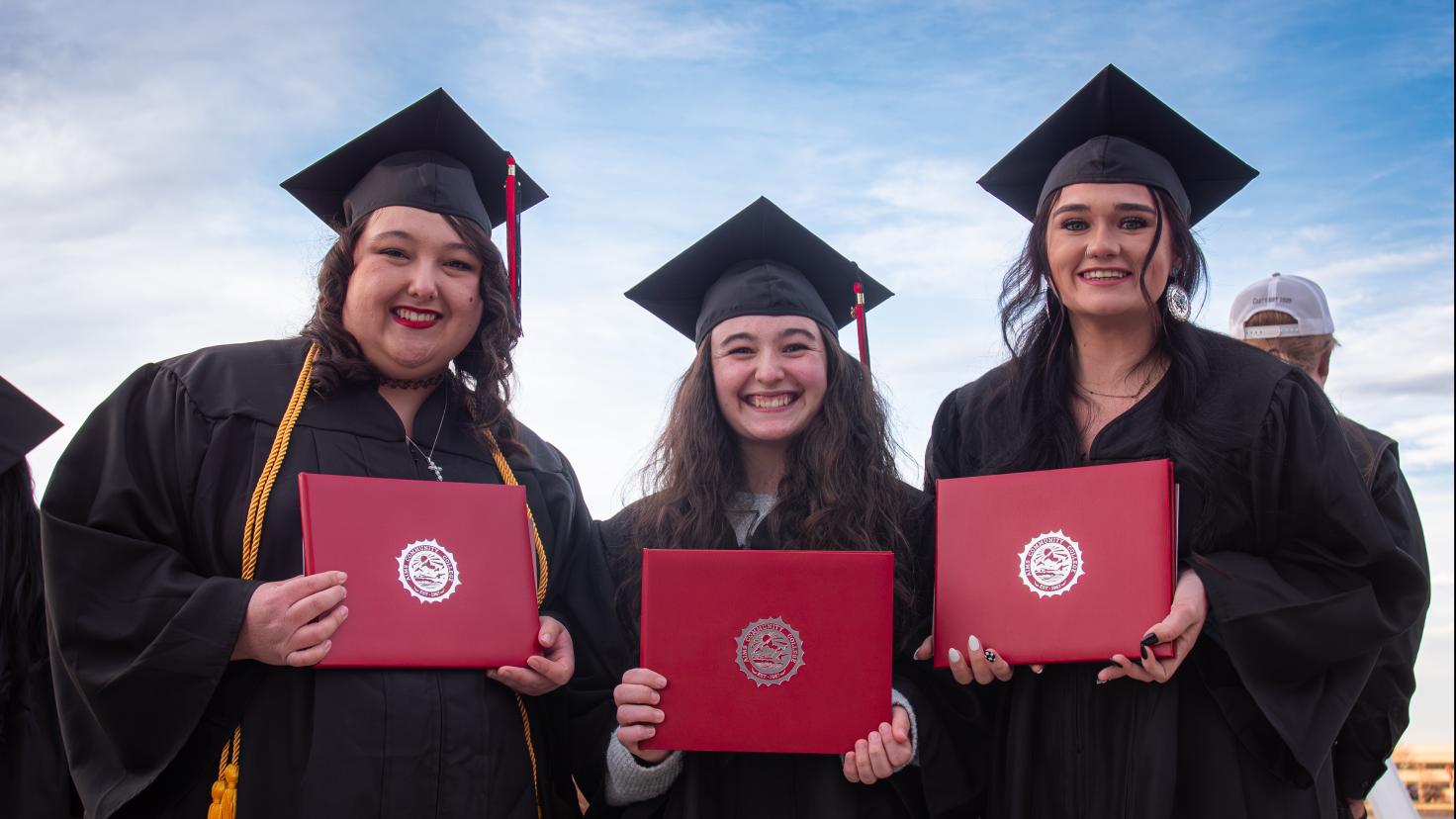 three graduates standing outdoors wearing caps and gowns holding diplomas