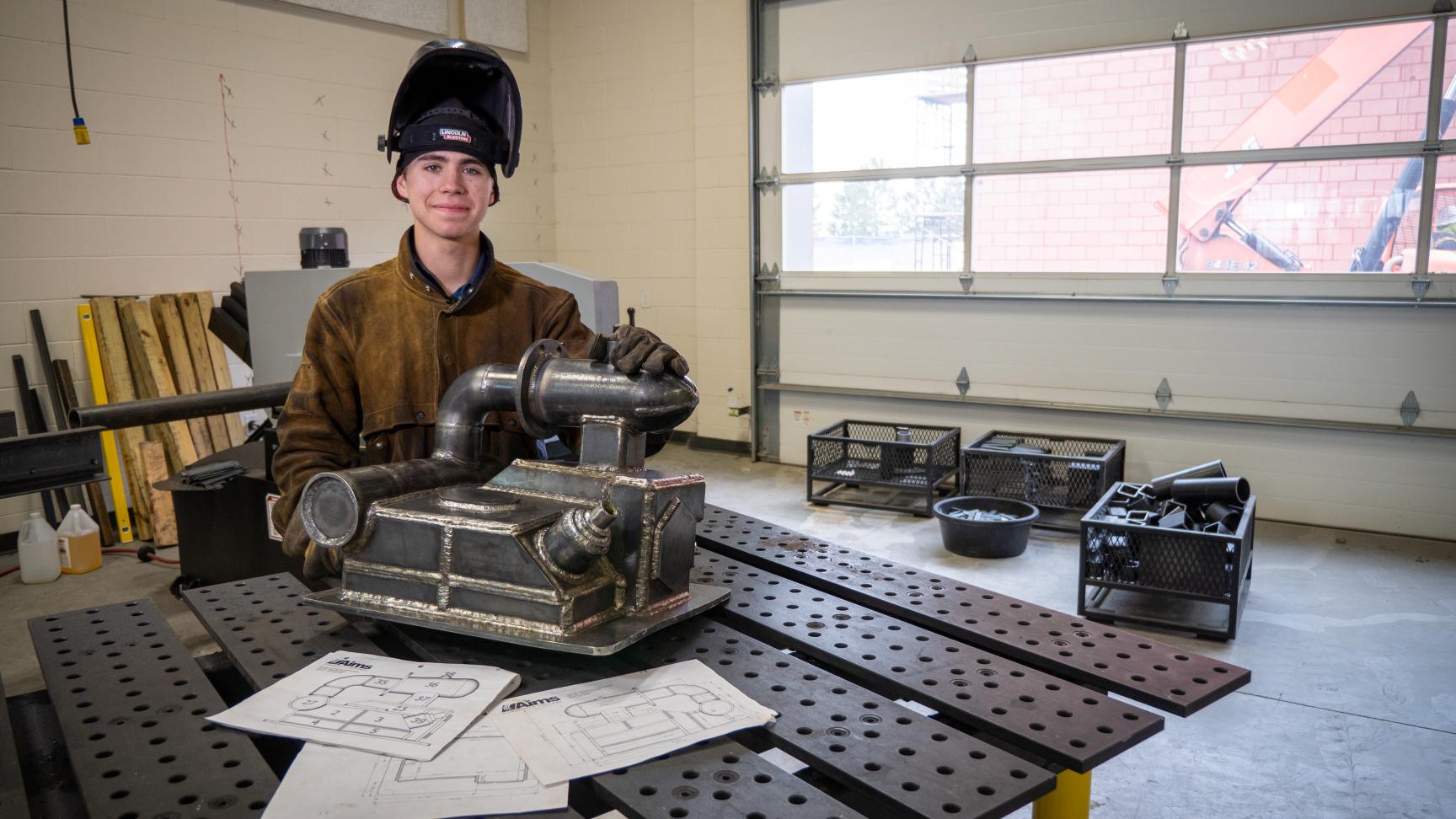A person in protective welding gear, stands smiling in front of a piece of welded metal