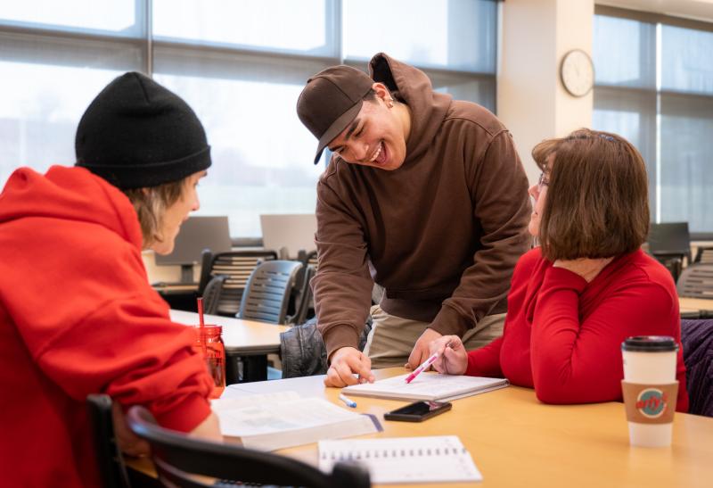 two students wearing red shirts sit across from each other at a table while a third student in brown hoodie leans over them to point at something on a notebook