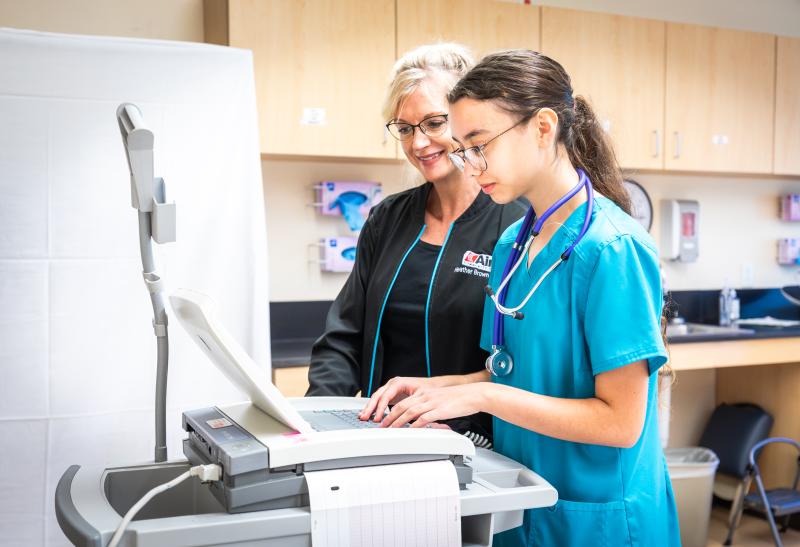 student with dark hair and ponytail in blue scrubs at computer station with teacher in dark scrubs next to her
