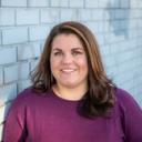 Smiling person with brown hair and purple shirt in front of a brick wall.