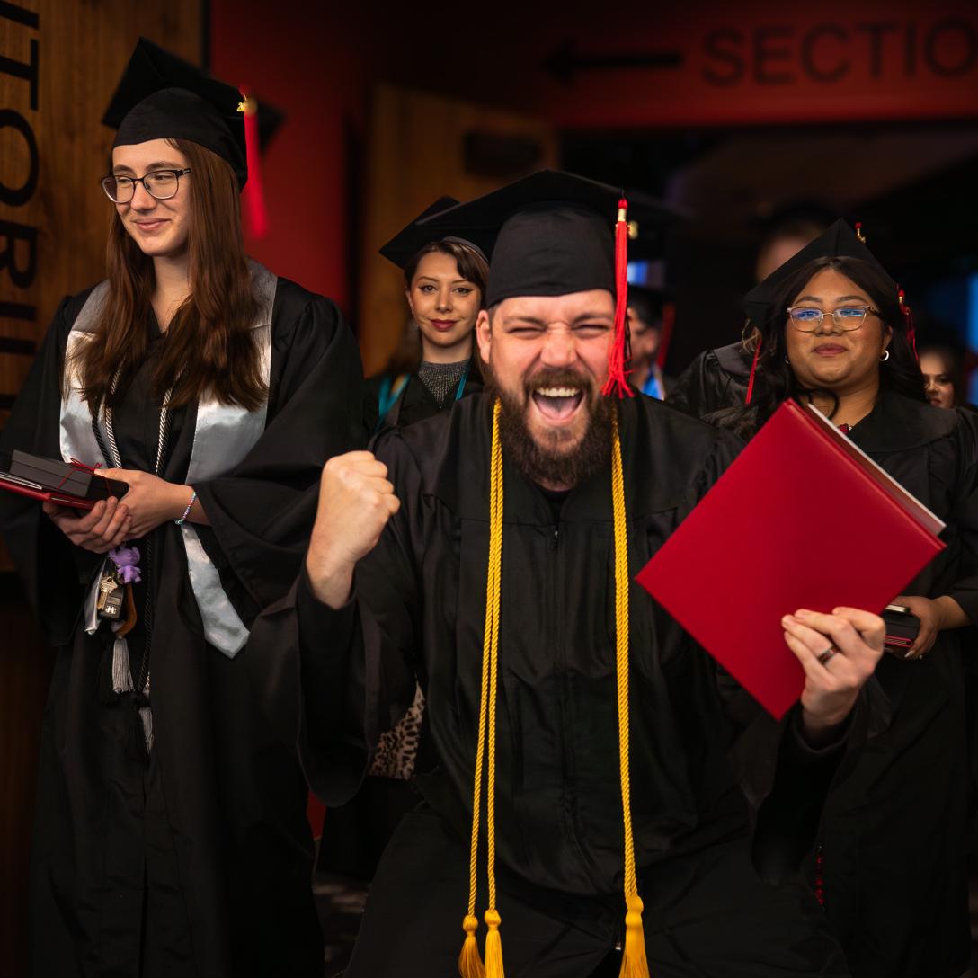 An Aims graduate looks at the camera with one hand raised in excitement and their diploma in the other hand