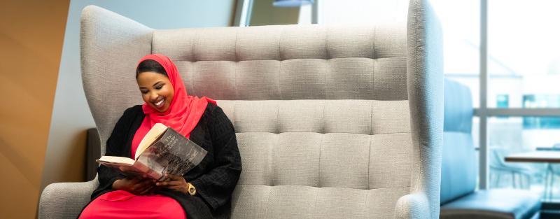A student sits in a large chair smiling and reading a book.