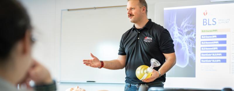 A person stands at the front of a classroom holding a CPR mask with a CPR practice dummy in front of them.