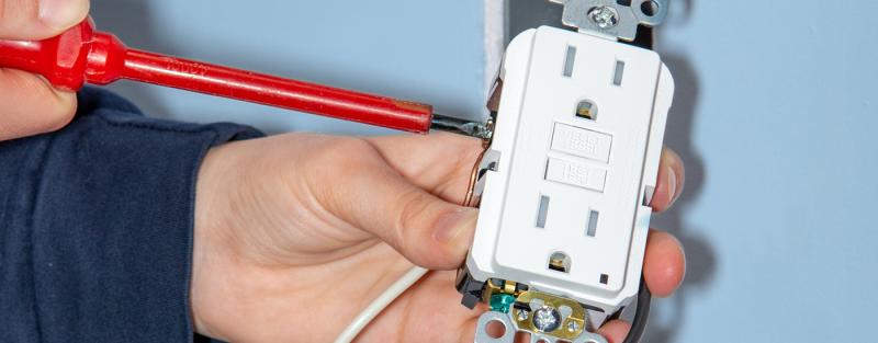 hand holding a white wall outlet with 2 plug spaces, using a red screwdriver to repair it
