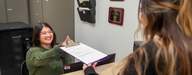 A person behind a desk in an office setting accepting a clipboard from a patient.