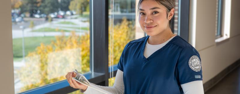 An Aims nursing degree student in dark blue scrubs holding a clipboard in class
