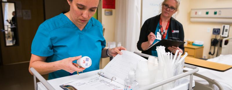 student wearing blue scrubs holds medicine bottle while standing in front of cart practicing administering medication while instructor watches