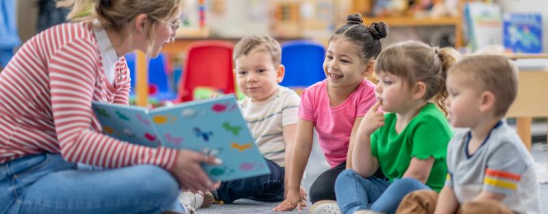 Three young children sitting cross-legged on floor with teacher reading to them from picture book