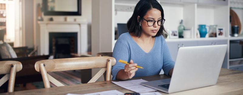 Woman wearing glasses sitting at table in home dining room working with papers and laptop