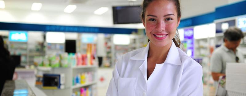 pharmacy technician in white lab coat stands arms crossed at the front of pharmacy