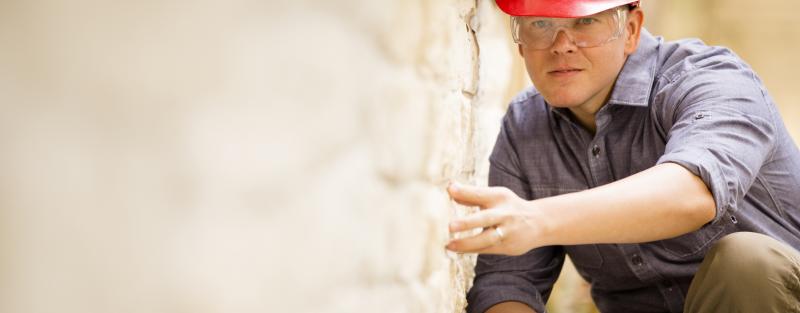 man wearing red hard hat crouches to inspect exterior wall of home