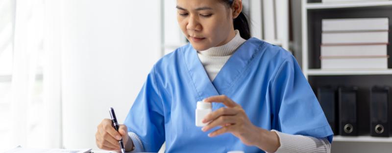 woman with brown hair wearing blue scrubs seated at table logging medicine information onto a sheet of paper