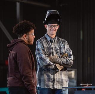 Two students stand talking. One is wearing a welding mask pushed up on their head and welding gloves.