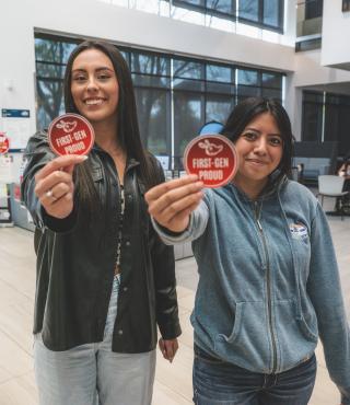 Two students stand holding First Gen proud stickers towards the camera
