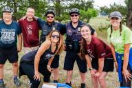  A group of people, including two police officers, are smiling and posing for a photo at an outdoor event. The group includes a mix of individuals wearing cycling gear, casual clothing, and Aims Community College attire. 