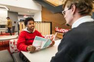 Student in a red sweatshirt holding a pamphlet, sitting in a modern lounge area speaking to an advisor