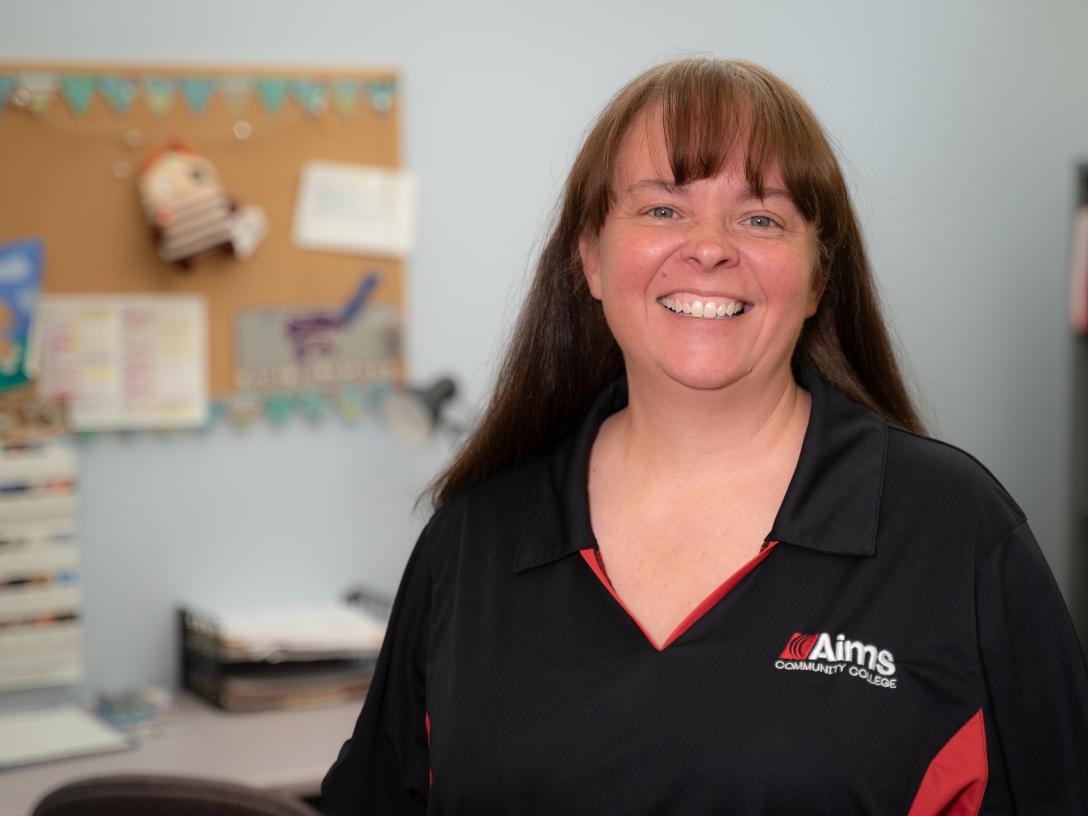 A woman with long brown hair stands smiling in an office wearing an Aims shirt. 