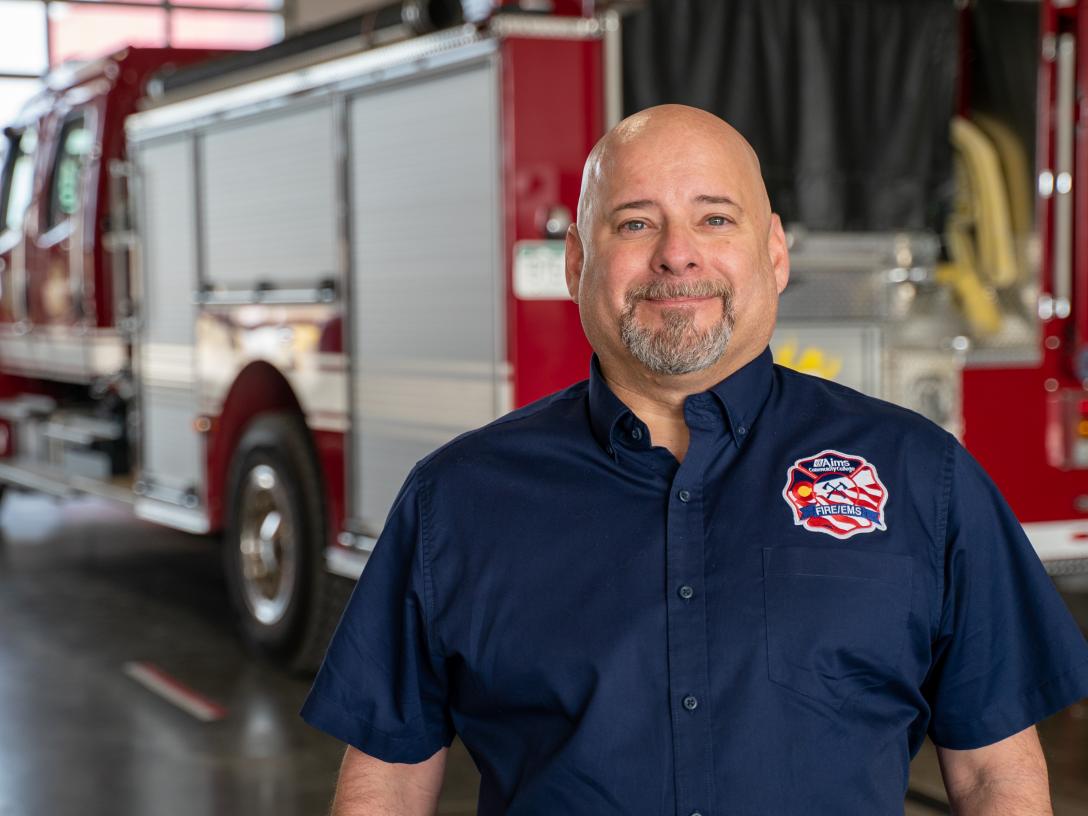 A firefighter stands smiling in front of a fire truck.