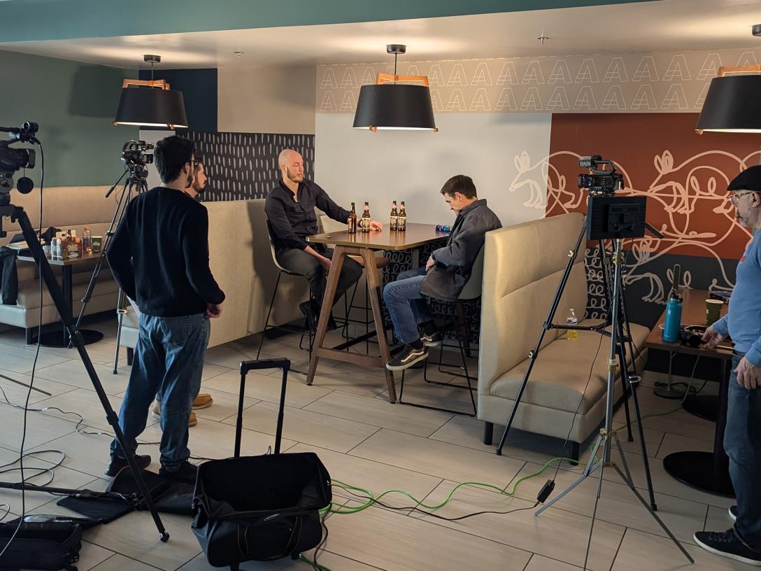 Two men at high top table talking while 3 men with film equipment record