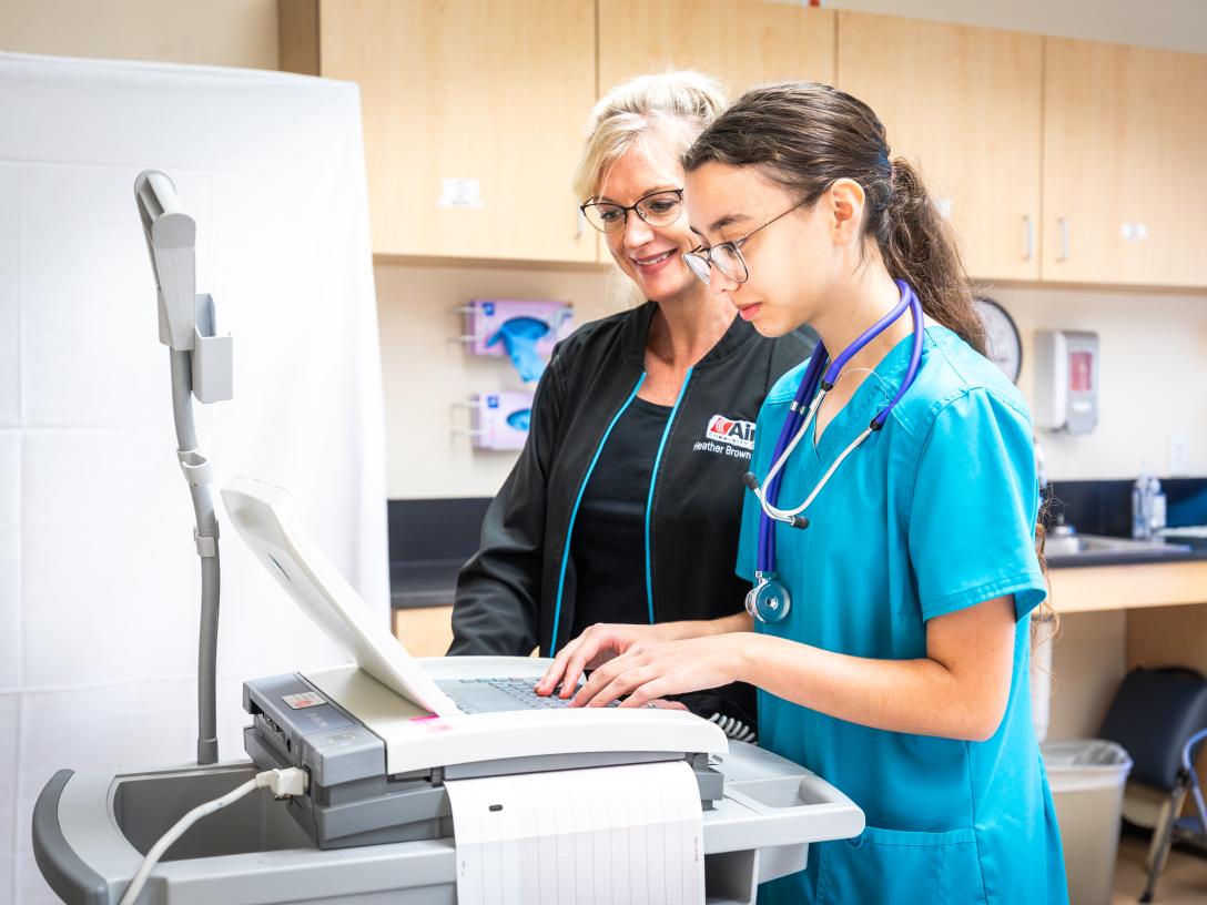 student with dark hair and ponytail in blue scrubs at computer station with teacher in dark scrubs next to her
