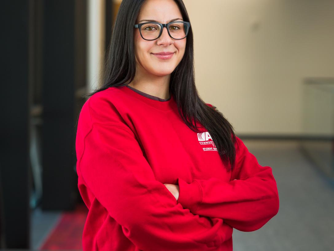 Aims student Katina Billiris standing with arms crossed in hallway, smiling, wearing red Aims sweatshirt