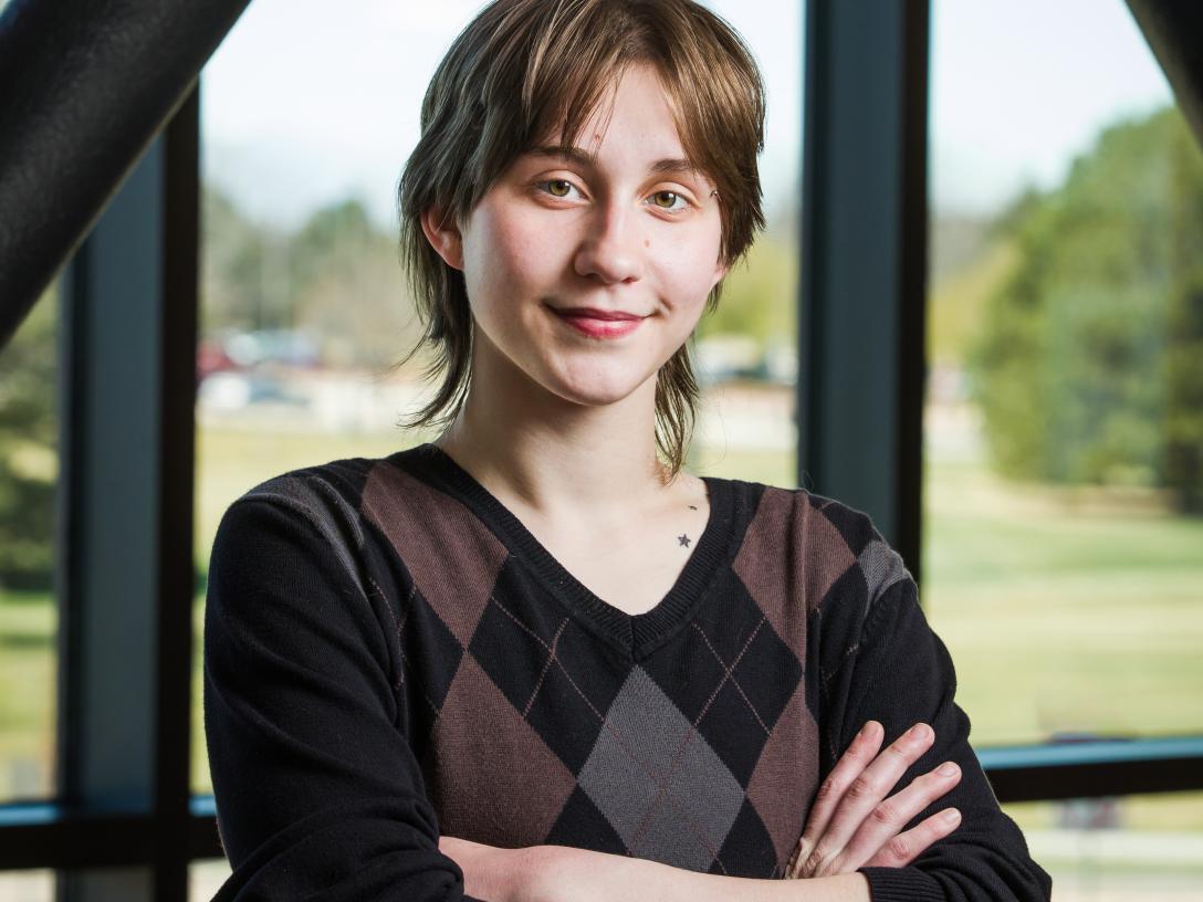 Aims student Onyx Hammond wearing black and brown argyle sweater with arms crossed in front of window overlooking campus