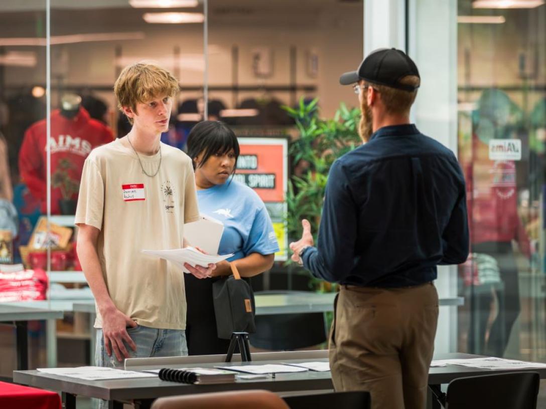 Student holding papers talks with an employer at a table during a campus job fair, with another student standing nearby