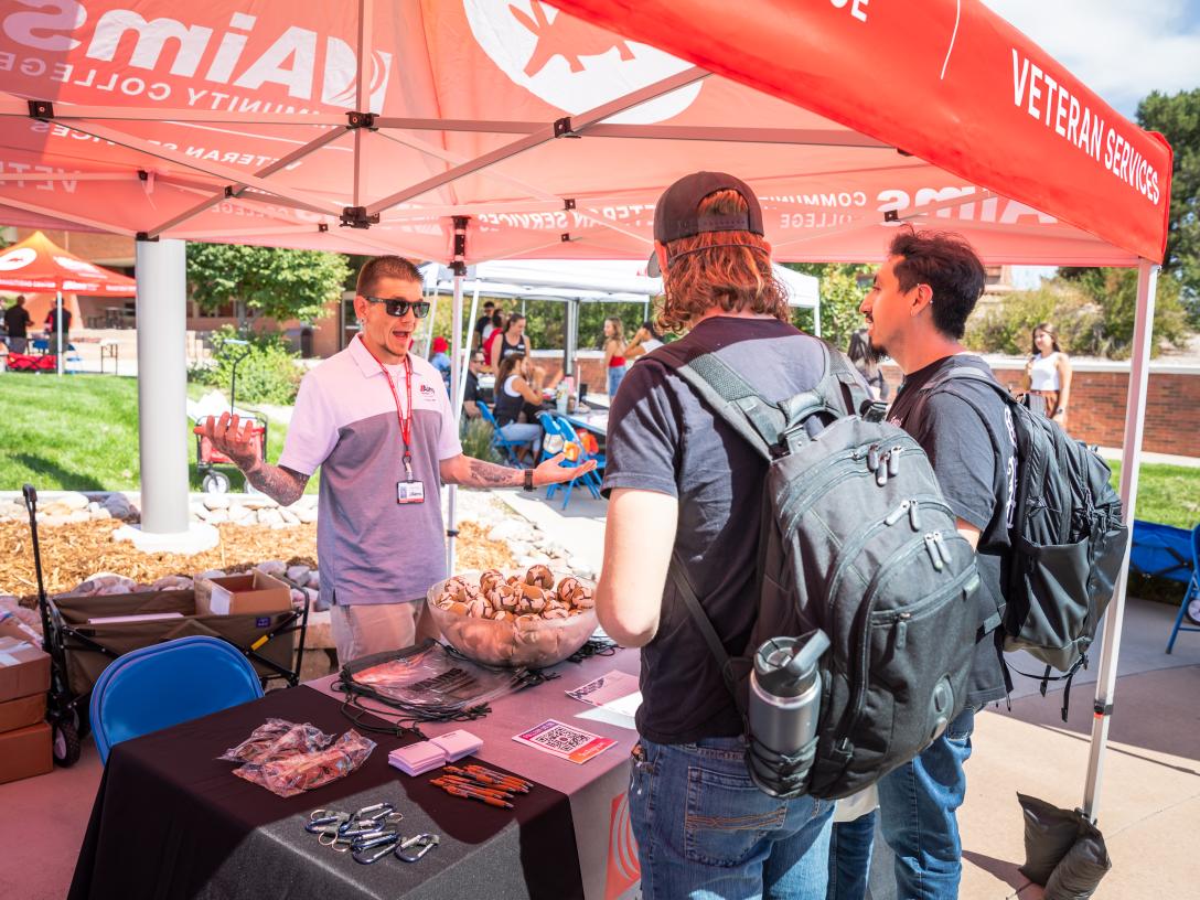 Veteran Services rep wearing Aims lanyard and sunglasses under Aims red tent talking to two students