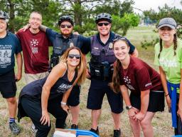  A group of people, including two police officers, are smiling and posing for a photo at an outdoor event. The group includes a mix of individuals wearing cycling gear, casual clothing, and Aims Community College attire. 