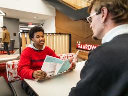 Student in a red sweatshirt holding a pamphlet, sitting in a modern lounge area speaking to an advisor