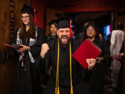 An Aims graduate looks at the camera with one hand raised in excitement and their diploma in the other hand