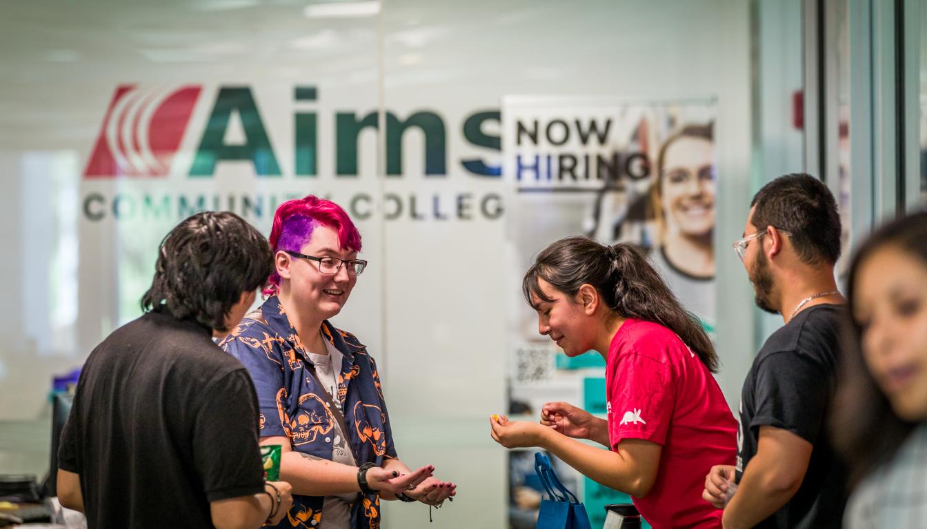 5 students standing in front of Aims Community College logo indoors talking and laughing