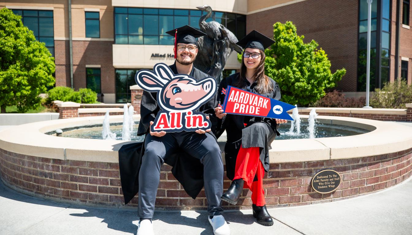 Students at Allied Health Building fountain with graduation caps and gowns holding signs