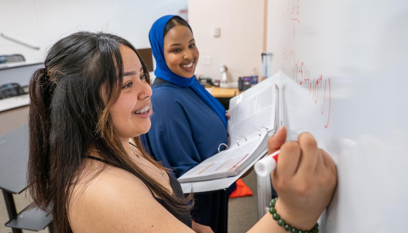 female students working on a math problem on a white board