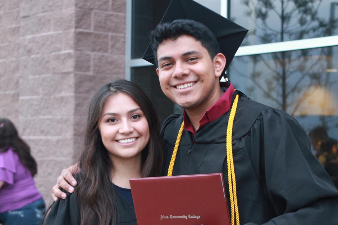 Two smiling Aims Community College students, with one wearing cap and gown and holding a diploma