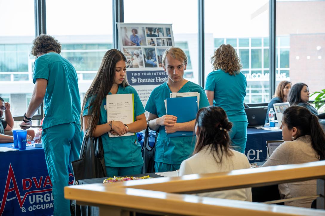 Aims students attend allied health career fair. Four students in blue scrubs talk with healthcare providers