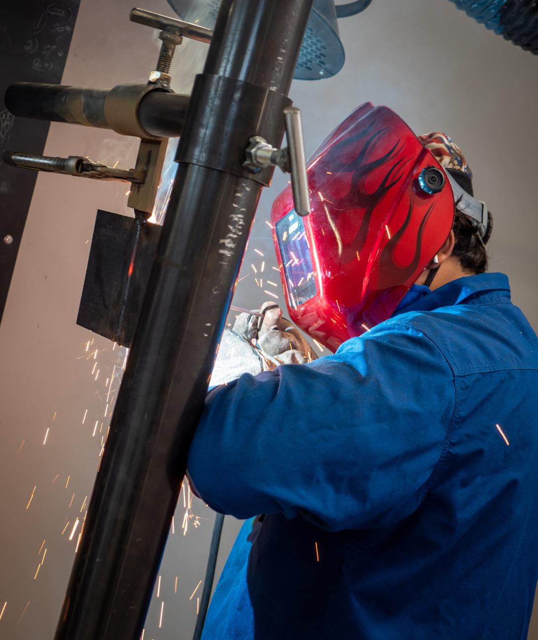 A student wearing a red welding mask decorated with flames welding metal as sparks fly