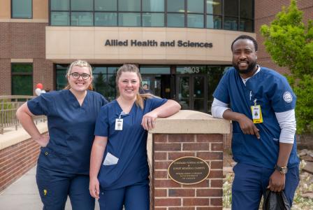 Three Aims Allied Health students in blue scrubs in front of Allied Health building